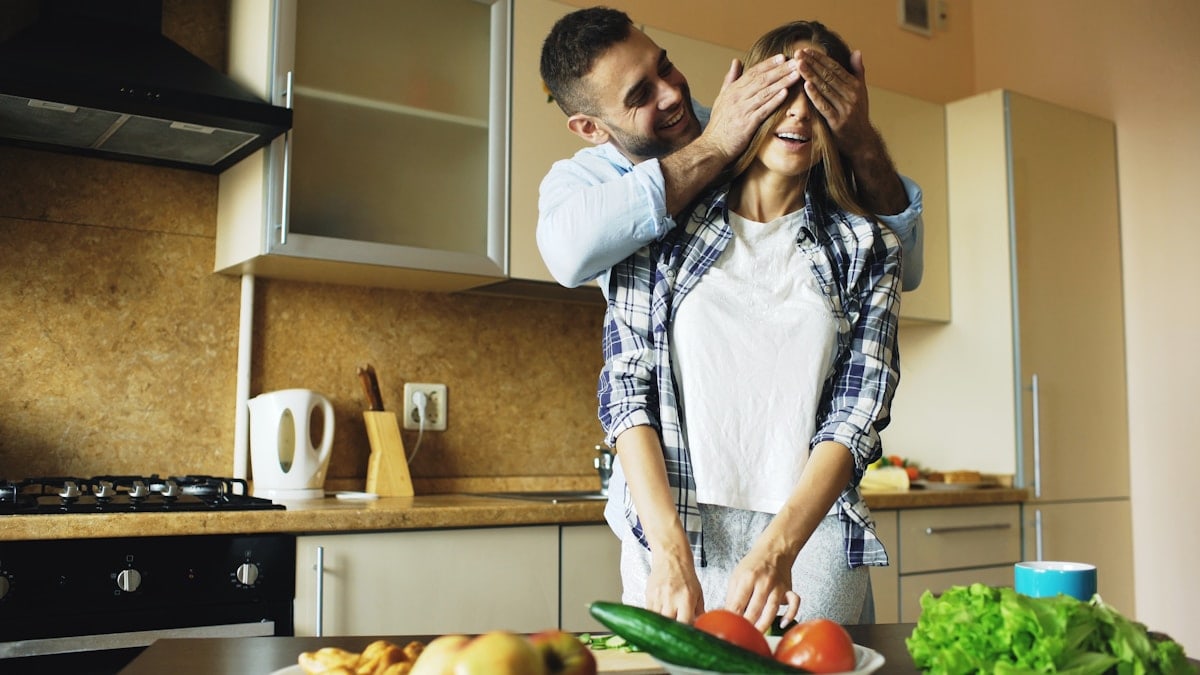 Young man covering girlfriends eyes with hands and surprising her in the kitchen at home. Loving couple cooking breakfast in the morning