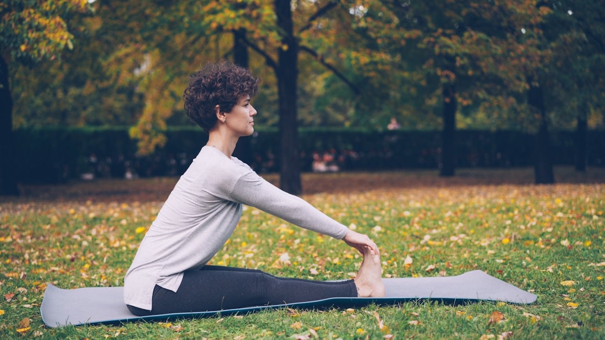Slim young lady is doing sequence of yoga asanas sitting on mat in park alone on warm autumn day with beautiful grass and trees around her. Recreation and sport concept.